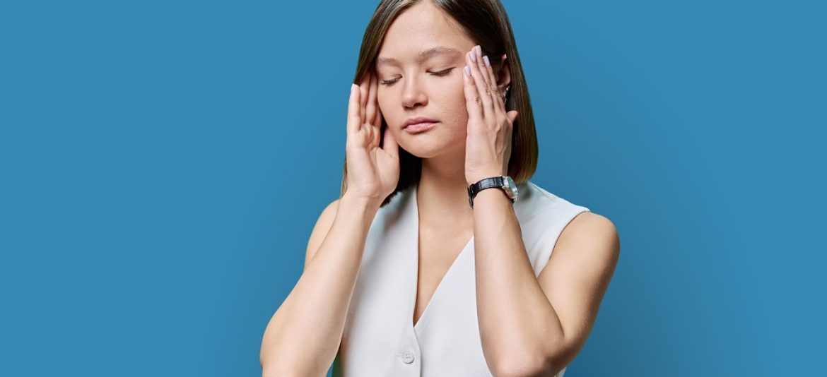 Young worried woman experiencing stress headache on blue background. Tired sad female holds hands on temples with eyes closed. Depression frustration migraine fatigue pain suffering, mental disorders